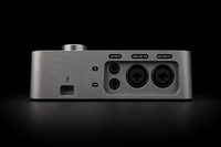 A silver metal audio interface device shown from the rear against a black background, featuring multiple audio and USB-C ports with labeled cutouts for outputs, microphone 2/3R, and microphone 1/6.