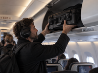 A man wearing black over-ear headphones and a black jacket places a black hard-sided luggage or equipment case into an overhead compartment inside an airplane cabin.