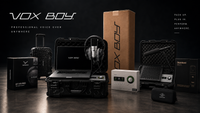 VOX BOY voice over kit displayed in a dark studio setup, including a laptop, rugged cases, headphones, a microphone, a small white audio device, a tall brown box, and branded boxes on a dark surface.