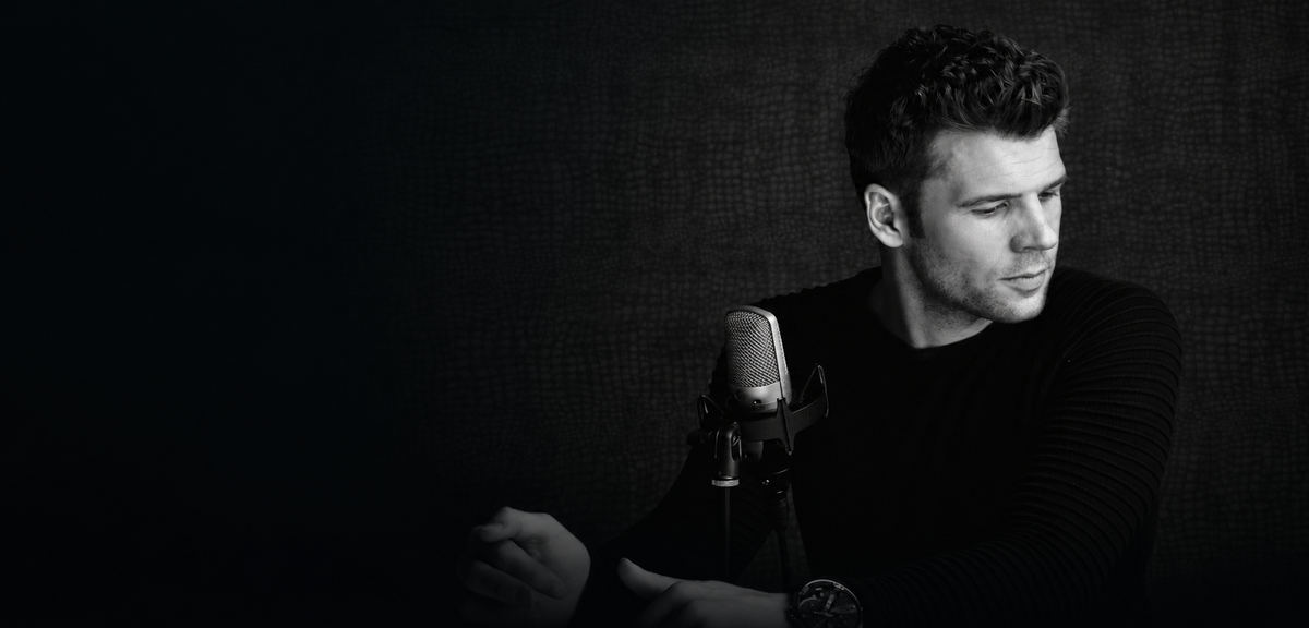 John Kubin beside a studio microphone, with a dark background